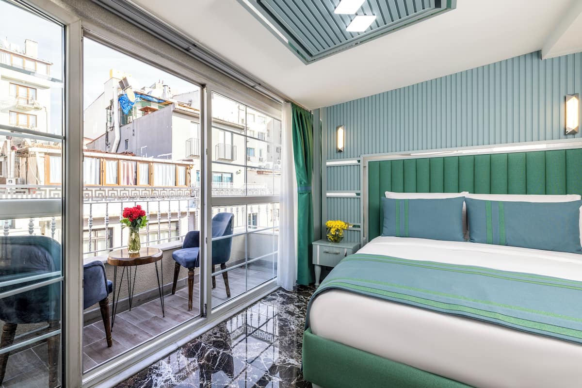 A hotel room with a large window, a balcony with chairs, a bed with green bedding, and a nightstand with a lamp.