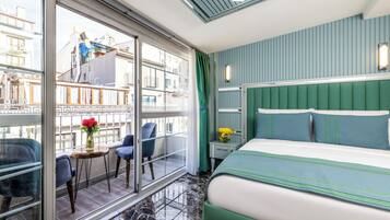 A hotel room with a large window, a balcony with chairs, a bed with green bedding, and a nightstand with a lamp.
