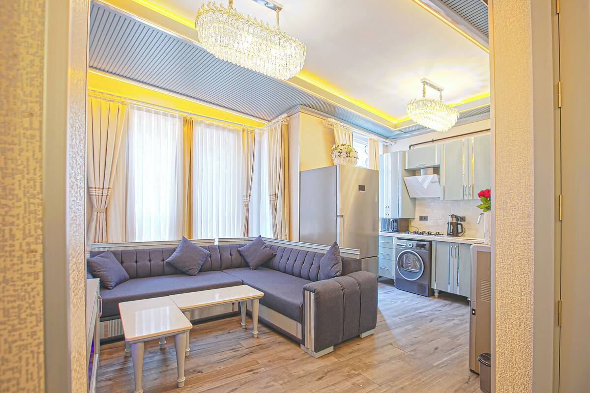 A modern living room with a sectional sofa, a small table, a washing machine, and a kitchen area.
