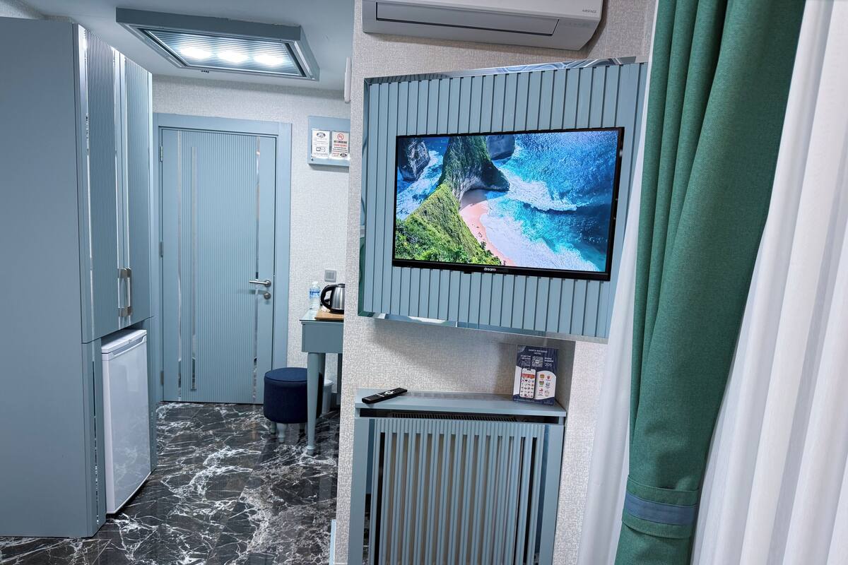 A hotel room with a TV on the wall, a minibar, a small table with a remote control, and a bathroom with a shower curtain.