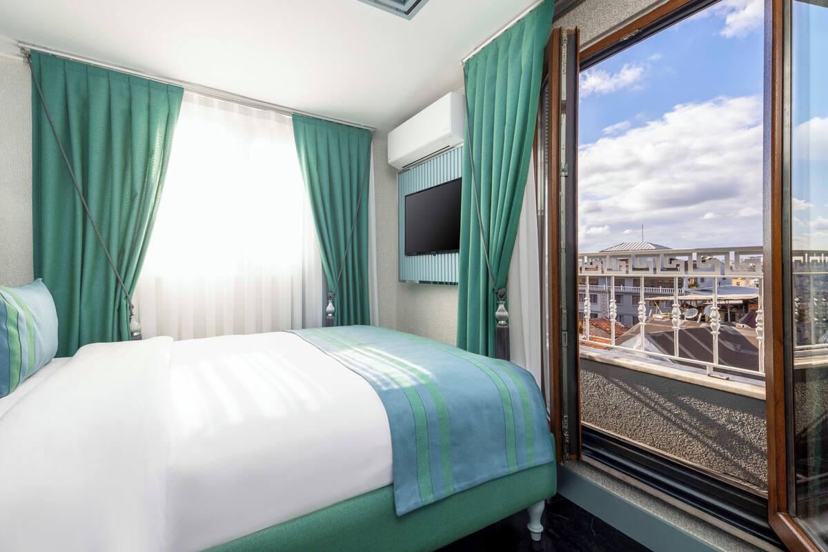 A hotel room with a bed, curtains, a TV, and a window with a view of buildings.