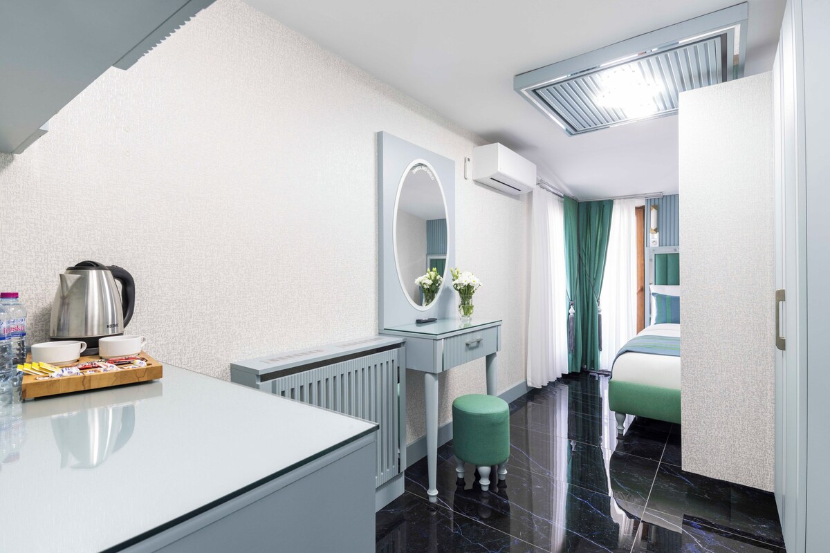 A modern hotel room with a white vanity, a mirror, a radiator, a green stool, and a bed with green curtains.
