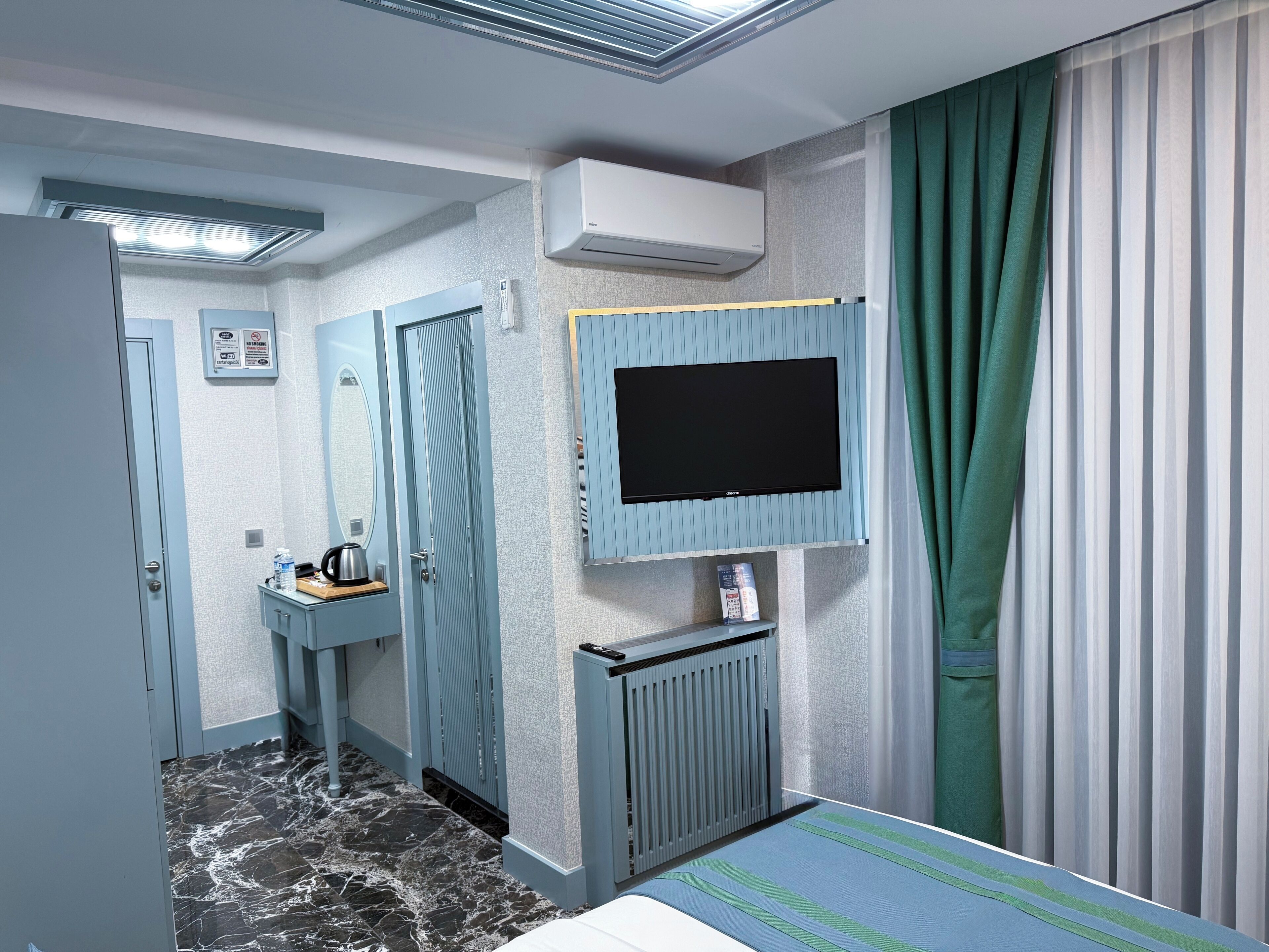 Habitació d'hotel amb un llit, una televisió penjada a la paret, un radiador, una taula petita amb un búl i un mirall.