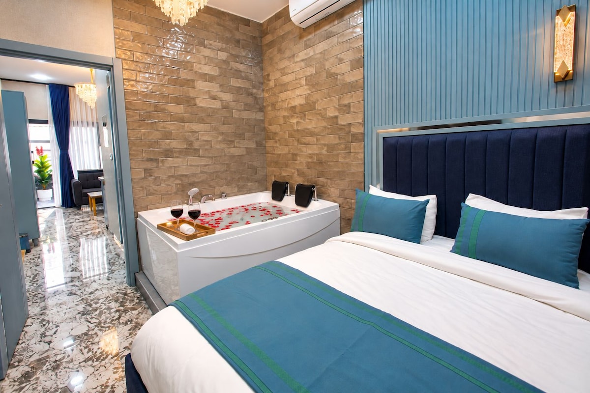A modern hotel room with a large bed, a bathtub with rose petals, and a view of the hallway.