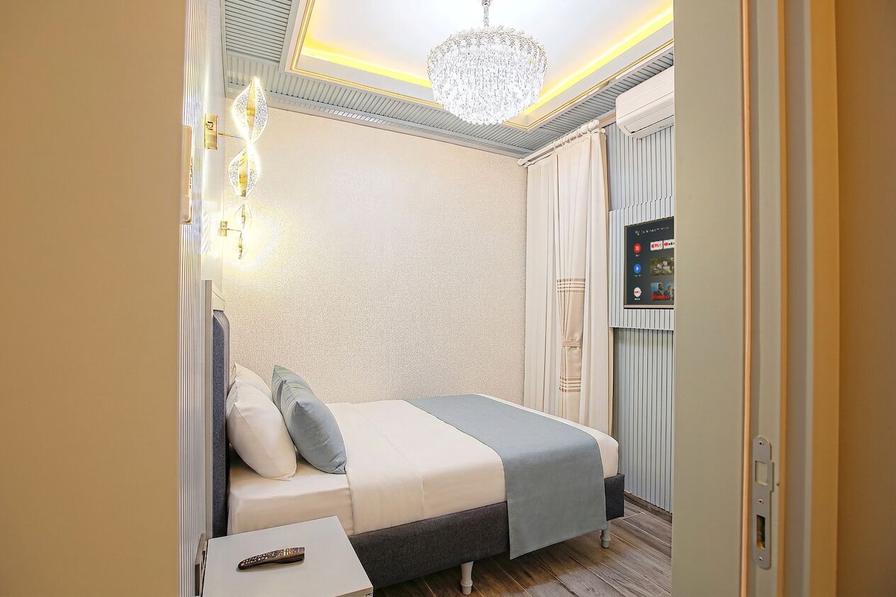 A hotel room with a bed, a TV, a chandelier, and a wall-mounted control panel.