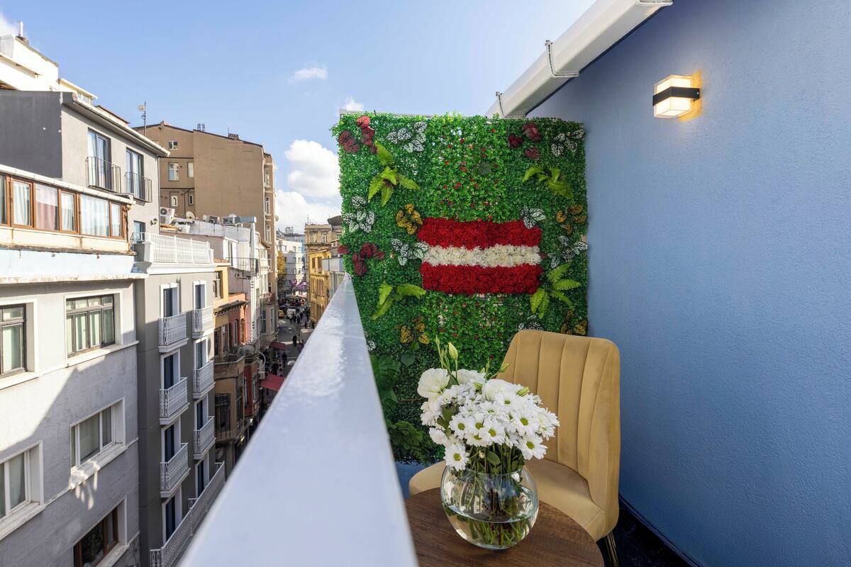 A balcony with a view of a cityscape, a decorative wall with a flag design, and a vase of white flowers on a table.