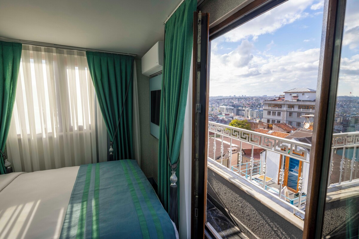 A hotel room with a bed, green curtains, and a window with a city view.