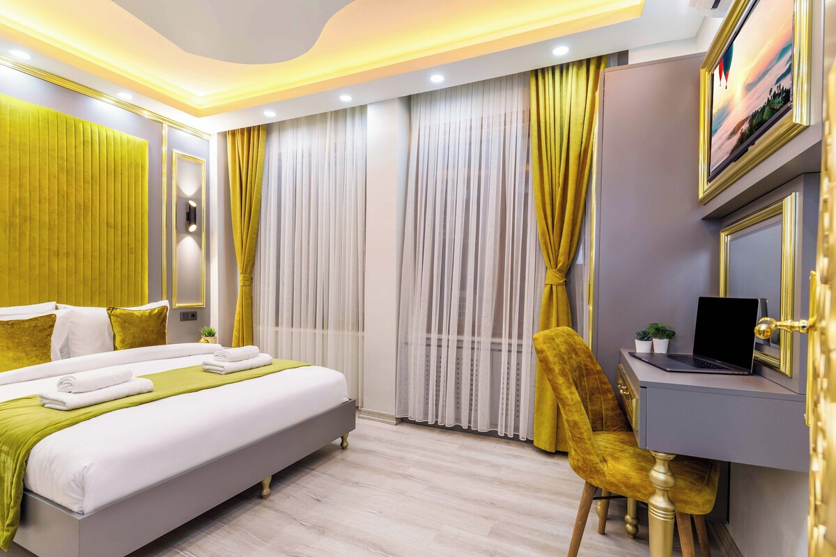 A modern hotel room with a large bed, a desk with a laptop, and a view of the city through the window.