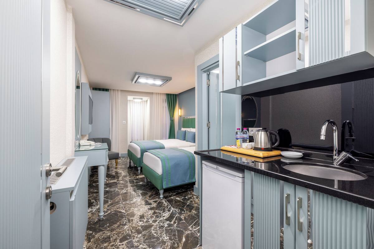 A compact hotel room with a kitchenette, a bed with green bedding, a small desk, and a bathroom with a toilet and sink.