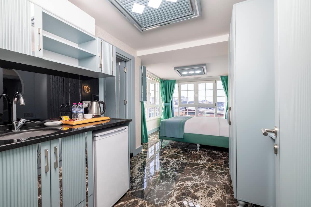 A compact hotel room with a kitchenette, a bed with a green bedspread, and a view of the city through the window.