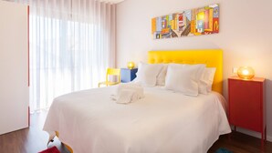 Een hotelkamer met een groot bed, witte beddengoed, een geel hoofdeinde, een kleurrijk kunstwerk aan de muur en een rode nachtkastje met een lamp.