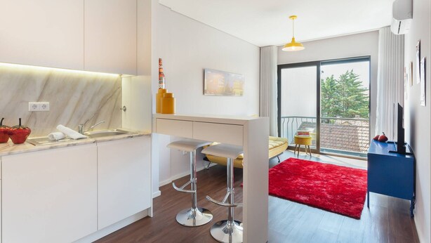 Een moderne keuken met witte kasten, een marmeren aanrechtblad en twee barkrukken. Er is een groot raam met uitzicht op bomen en een balkon.