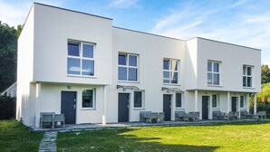Un edificio moderno de dos pisos con techo plano, ventanas grandes y un pequeño jardín delantero con mobiliario exterior.