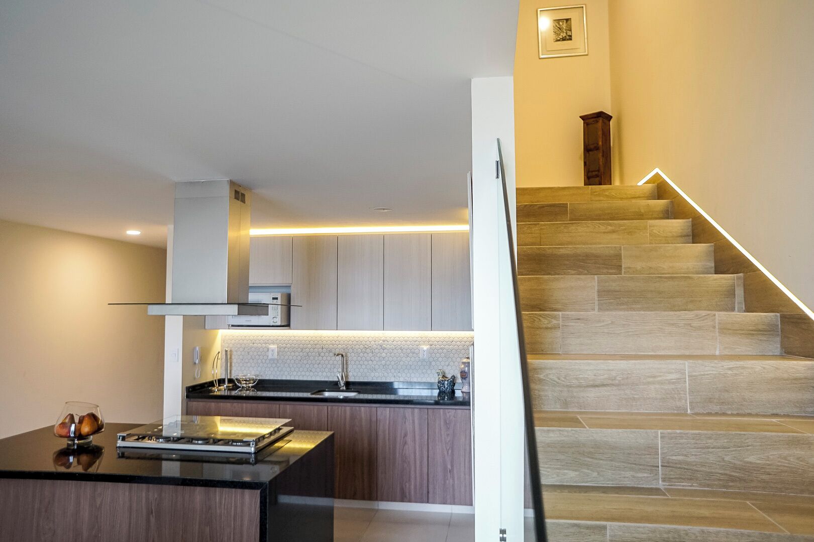Una cocina moderna con una isla central, horno empotrado y pileta de acero inoxidable. Se ve una escalera con escalones de madera y una lámpara a la derecha.