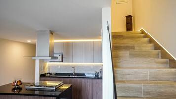 Una cocina moderna con una isla central, horno empotrado y pileta de acero inoxidable. Se ve una escalera con escalones de madera y una lámpara a la derecha.