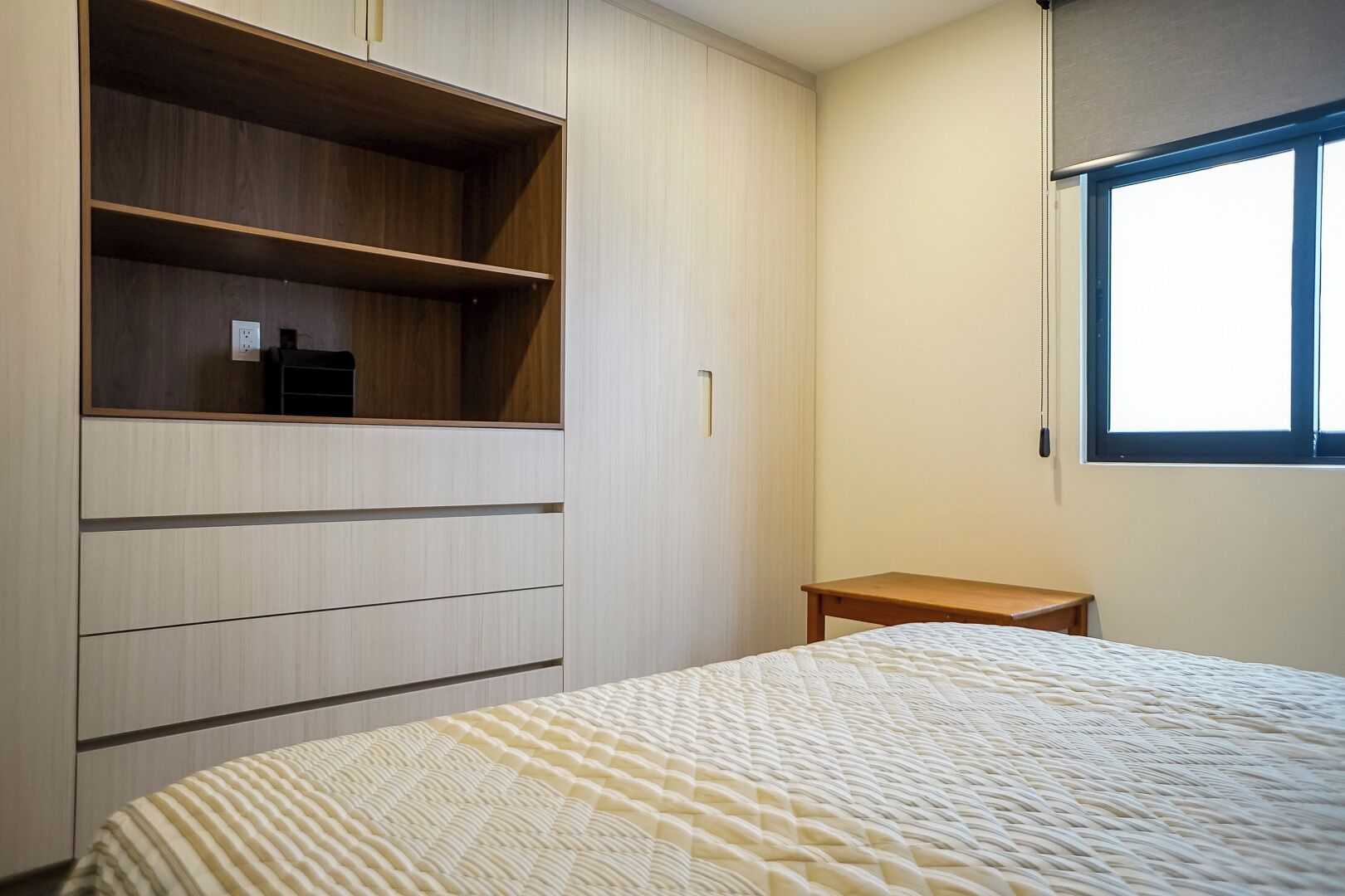 Une chambre à coucher comprenant un lit, une armoire en bois avec plusieurs tiroirs, une petite table de chevet en bois et une fenêtre avec des stores.