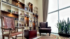 A living room with a wooden rocking chair, a dark armchair, a bookshelf, and a large window with curtains.
