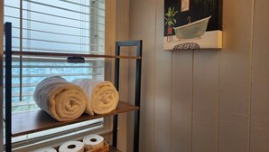 Un bagno con un porta asciugamani con due asciugamani arrotolati, un cesto con carta igienica e uno scaffale a parete con una pianta in vaso e un quadro.