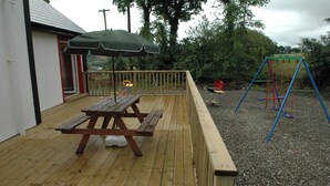 A wooden deck with a picnic table and an umbrella, a swing set, and a gravel area. - Willowfarmhouse Accommodation (Milford)