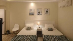 A hotel room with two beds, a bedside table with a lamp, a chair, and wall art. - Maria Pyramids House - Standard Double or Twin Room (Giza City)