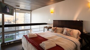 Una habitación de hotel moderna con una cama grande, un televisor montado en la pared, un balcón con vista a la ciudad y una pequeña mesa con una lámpara.