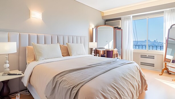 Hotellihuone, jossa on suuri sänky, yöpöytä, peili ja ikkuna, josta avautuu näkymä kaupunkiin.