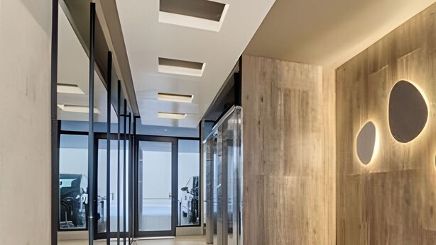 Un intérieur moderne avec un long couloir, des portes vitrées et un mur en bois agrémenté d’un éclairage circulaire.