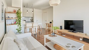 Un salon moderne avec un canapé, une table basse et un téléviseur. Au fond, on aperçoit une cuisine avec un coin repas.