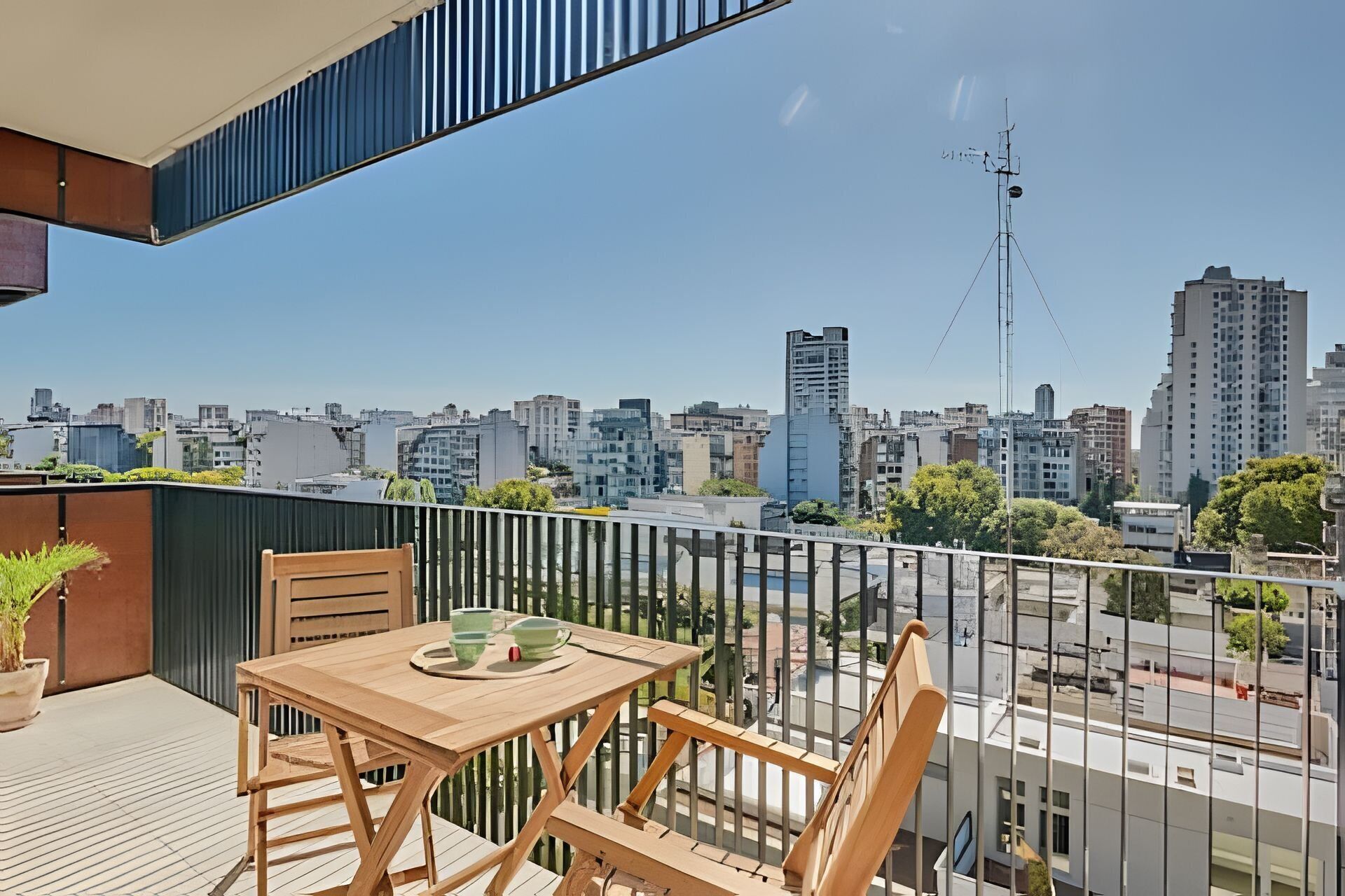 Een balkon met een houten tafel en stoelen, met uitzicht op een stadsbeeld met verschillende gebouwen en een hoge zendmast.