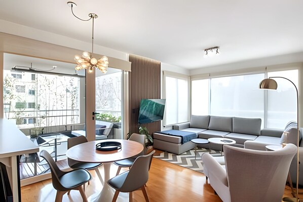 A modern living room with a round dining table, grey sofa, and a large window offering a city view. - Great 2BR with Heated Rooftop Pool, Gym & Hot Tub (Buenos Aires)