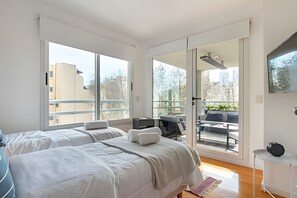 A modern bedroom with two beds, a balcony with a table and chairs, and large windows. - Great 2BR with Heated Rooftop Pool, Gym & Hot Tub (Buenos Aires)