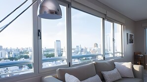 A modern living room with a large window offering a city view, a sofa with cushions, and a pendant light. - Luxury 2BR Apartment with Pool, Gym & Spa (Buenos Aires)