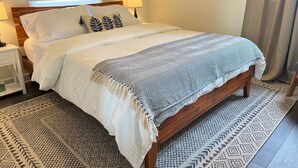 Una cama bien hecha con un plaid estampado, una lámpara de noche y un tapete estampado sobre un piso de madera.