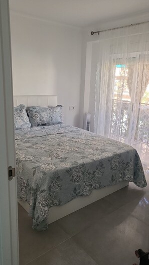 A bedroom with a large bed, a white headboard, and a view of the outdoors through sheer curtains.