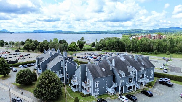 A residential complex with multiple two-story buildings, a parking lot, and a view of a lake and mountains in the background. - Condotel (Magog)