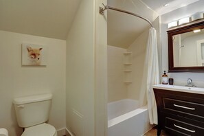 A bathroom with a white toilet, a bathtub, a shower curtain, a wooden vanity with drawers, a mirror, and a framed picture on the wall.