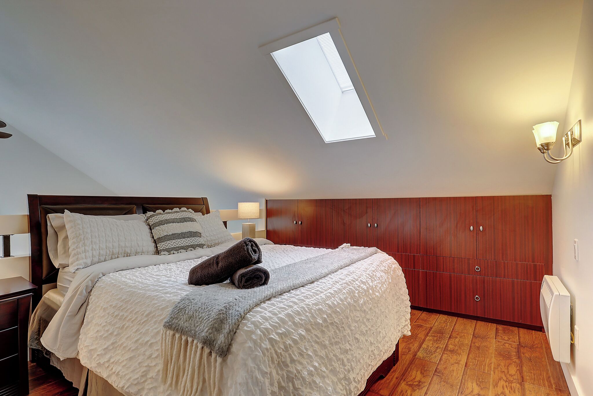 A bedroom with a skylight, a bed with a white duvet and pillows, a wooden wardrobe, and a bedside lamp.