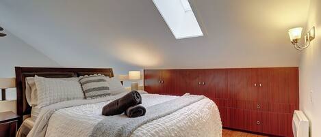 A bedroom with a skylight, a bed with a white duvet and pillows, a wooden wardrobe, and a bedside lamp.