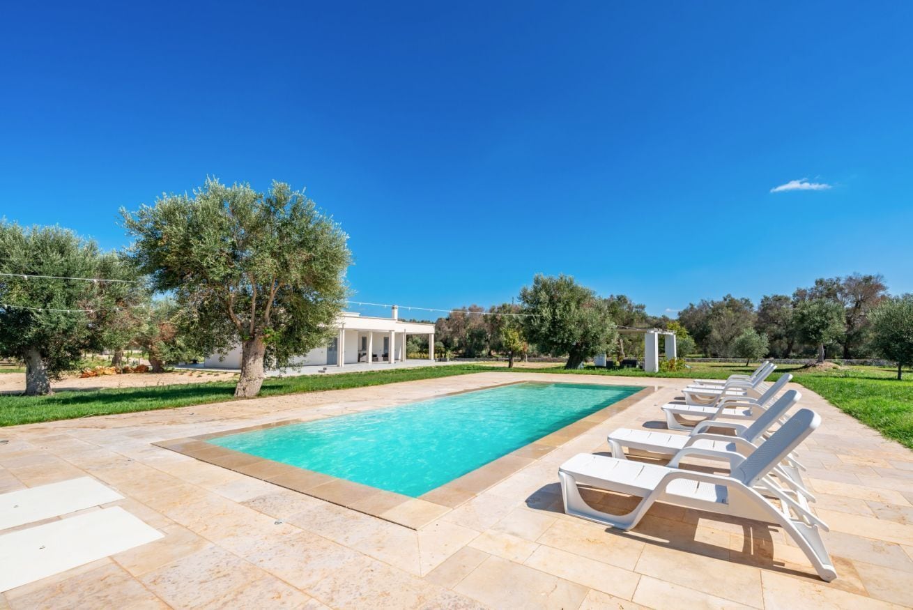 A rectangular swimming pool with lounge chairs on one side, surrounded by a paved deck and trees.
