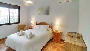 A neatly made bed with white linens and towels, a wooden headboard, two bedside tables with lamps, a wicker chair, and a small table with a vase. - Cómpeta Rural - Casa La Niña by Ruralidays (Málaga)
