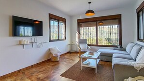 A modern living room with a sofa, a coffee table, a TV mounted on the wall, and large windows with blinds. - Cómpeta Rural - Casa La Niña by Ruralidays (Málaga)