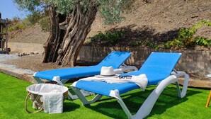 Two sun loungers with white frames and blue cushions, a white bucket, and a wicker basket on a well-maintained lawn with a tree and a stone wall in the background. - Cómpeta Rural - Casa La Niña by Ruralidays (Málaga)