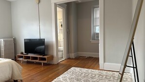 Ein Schlafzimmer mit Fernseher auf einem Holzboden, einem Heizkörper, einem weißen Teppich und einem Fenster mit Jalousien.