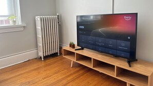 Ein modernes Wohnzimmer mit einem Flachbildfernseher auf einem Holzständer, einer Heizung und einem Fenster mit Jalousien.