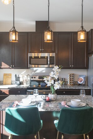 Una cocina moderna con armarios de madera oscura, electrodomésticos de acero inoxidable y encimera de mármol. Hay dos sillas verdes y una mesa de comedor preparada para dos personas.