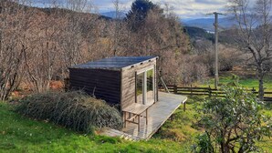 A small wooden cabin with a sloped roof, surrounded by a wooden deck, set in a lush green landscape with trees and hills in the background.