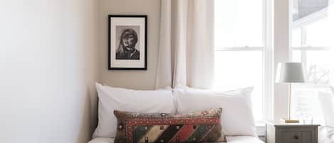 Ein ordentlich bezogenes Bett mit weißen und gemusterten Kissen, ein gerahmtes Bild an der Wand und ein Nachttisch mit Lampe und Glas.