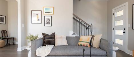 A living room with a grey sofa, a coffee table with books and a board game, a staircase, and a door with windows.