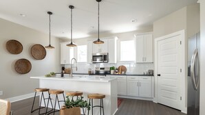 Een moderne keuken met witte kasten, een centraal eiland met krukken, hangende pendellampen en een roestvrijstalen koelkast.