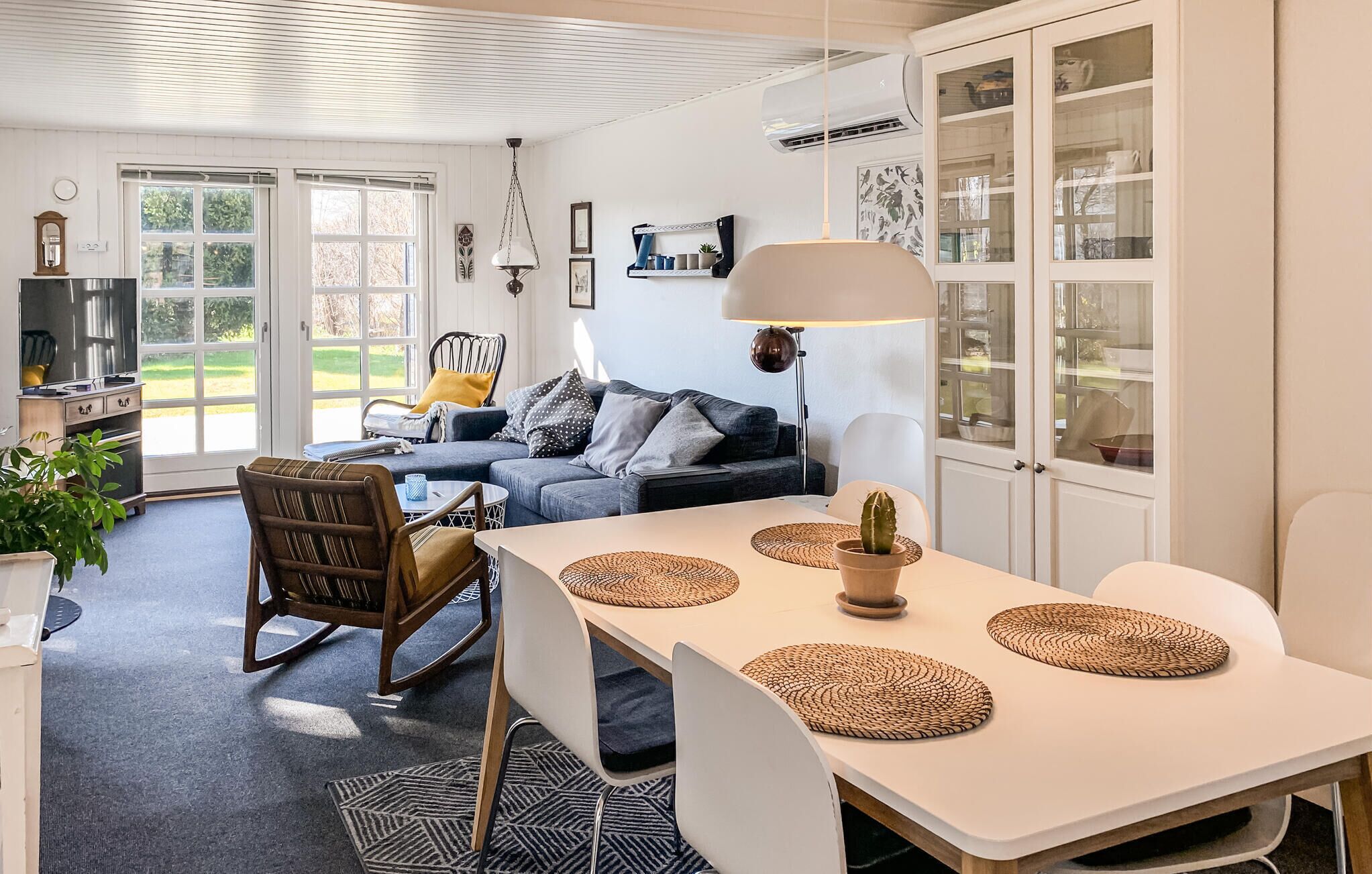 A modern kitchen with a dining area, featuring a white table with woven placemats, a sofa with cushions, and a hanging lamp.